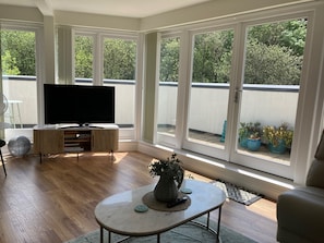 Uma sala de estar ampla com uma televisão grande, piso de madeira, uma mesa de centro redonda e janelas grandes com vista para o exterior.