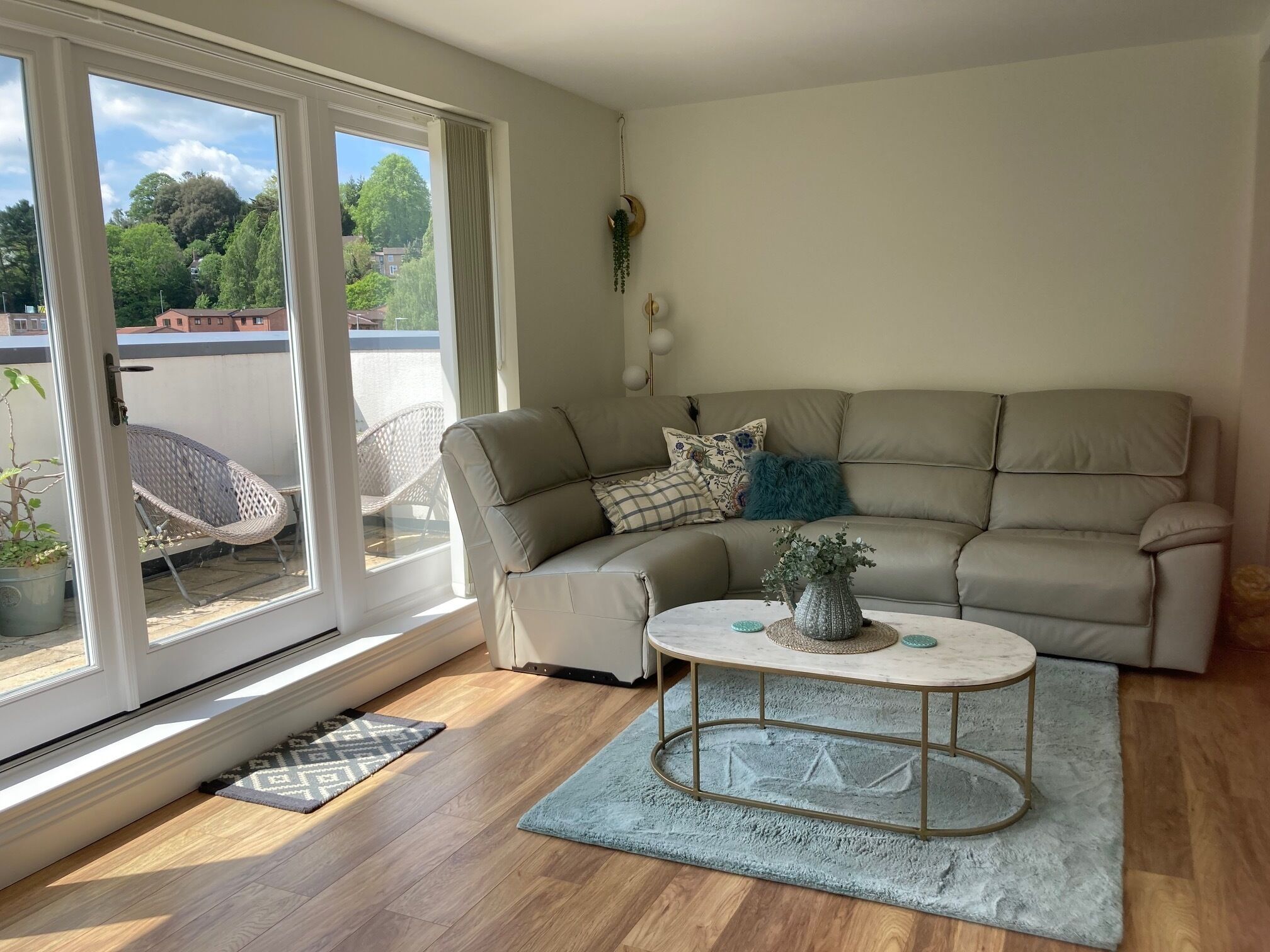 Een woonkamer met een beige bank, een ronde salontafel en een schuifdeur naar een buitenruimte met een stoel en een bloempot.