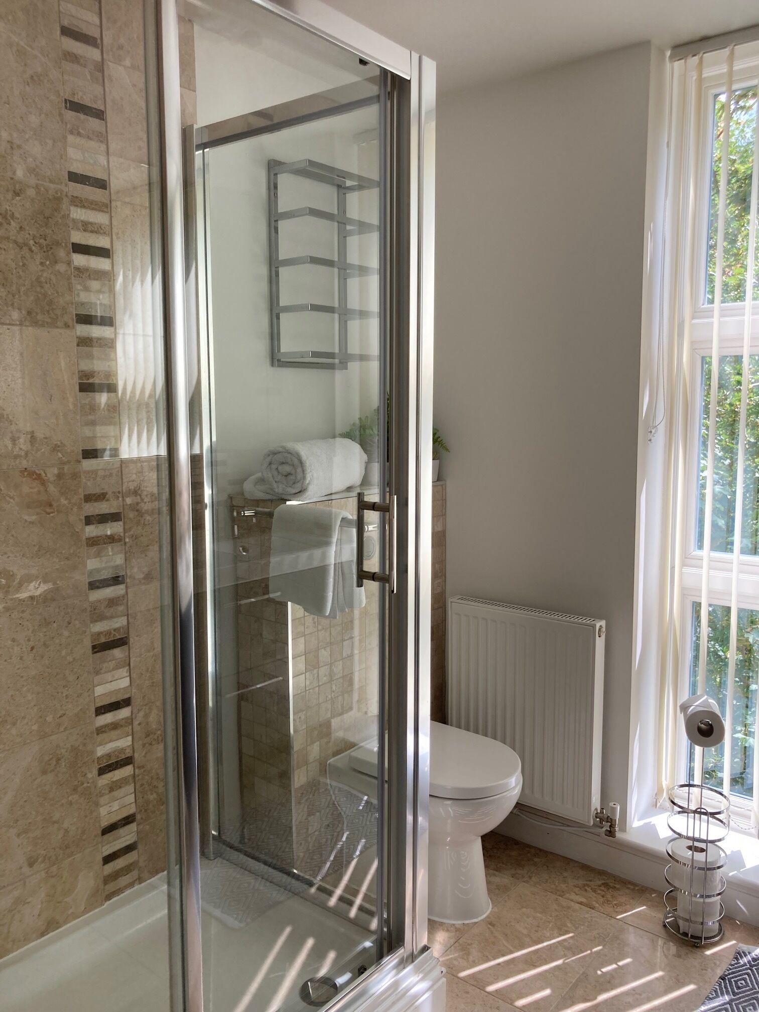 A bathroom with a glass shower enclosure, a toilet, a radiator, and a window with curtains.