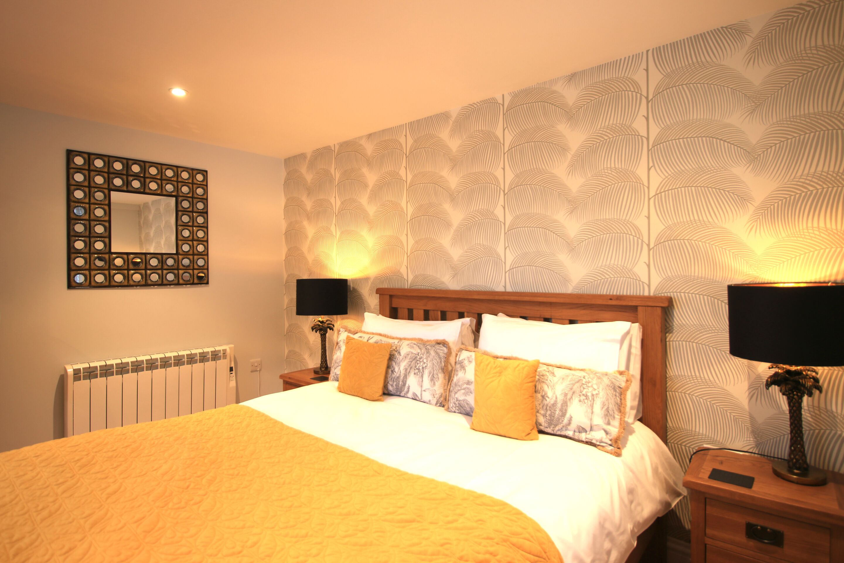 Quarto de hotel com cama, duas luminárias de cabeceira, um espelho e papel de parede estampado.