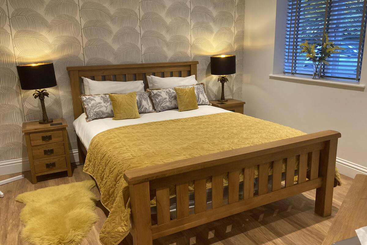 Una cama bien hecha con cabecera de madera, dos mesitas de noche con lámparas, una alfombra mullida y una ventana con persianas.