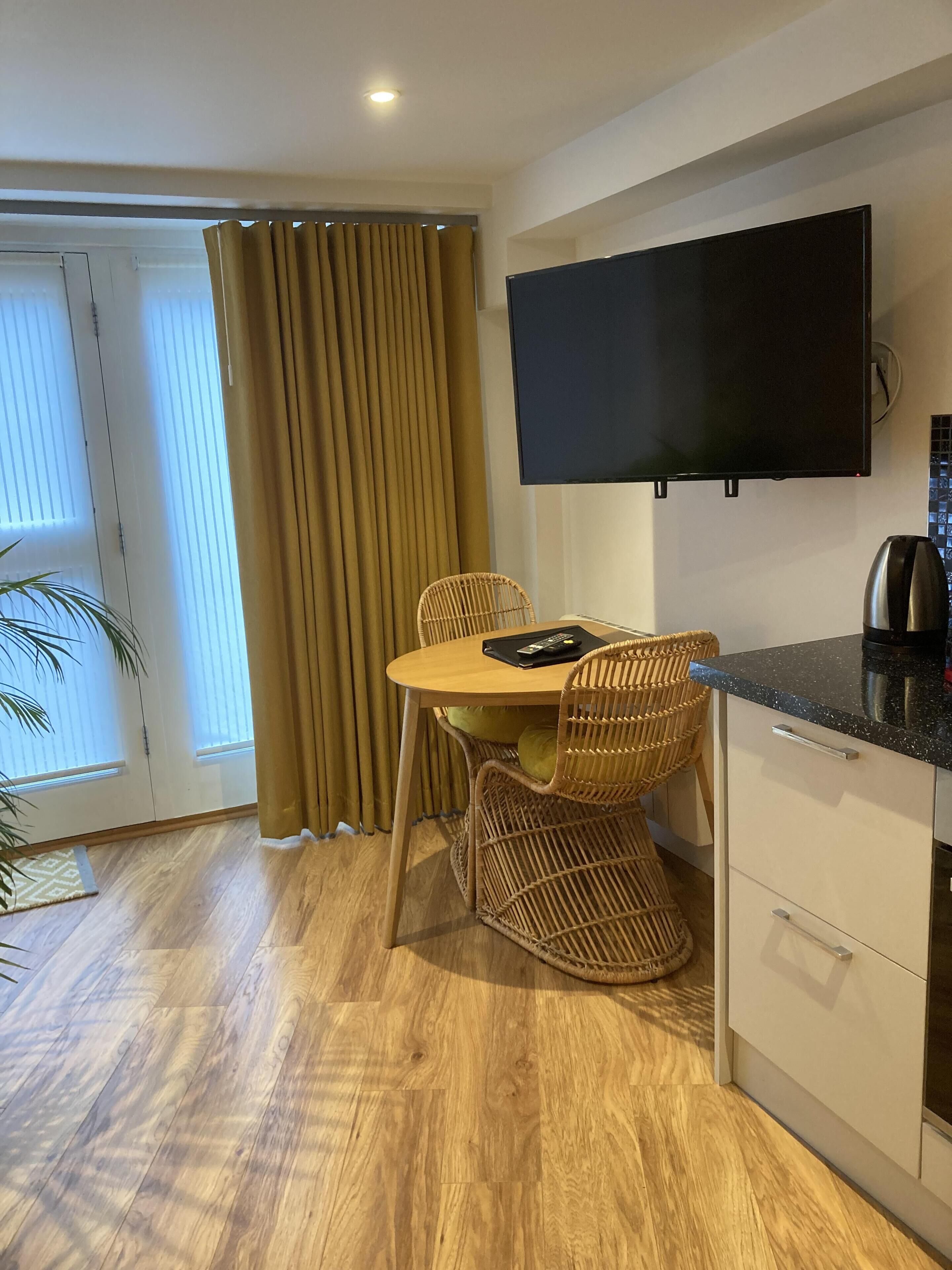 A modern room with a wooden floor, a dining table with chairs, a flat-screen TV mounted on the wall, and a kitchen area with a coffee maker.