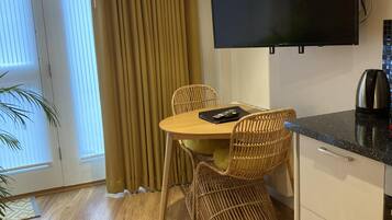 A modern room with a wooden floor, a dining table with chairs, a flat-screen TV mounted on the wall, and a kitchen area with a coffee maker.