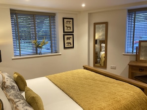 A bedroom with a bed, a bedside table, a mirror, and a window with blinds. - Streamside Apartments (Yeovil)