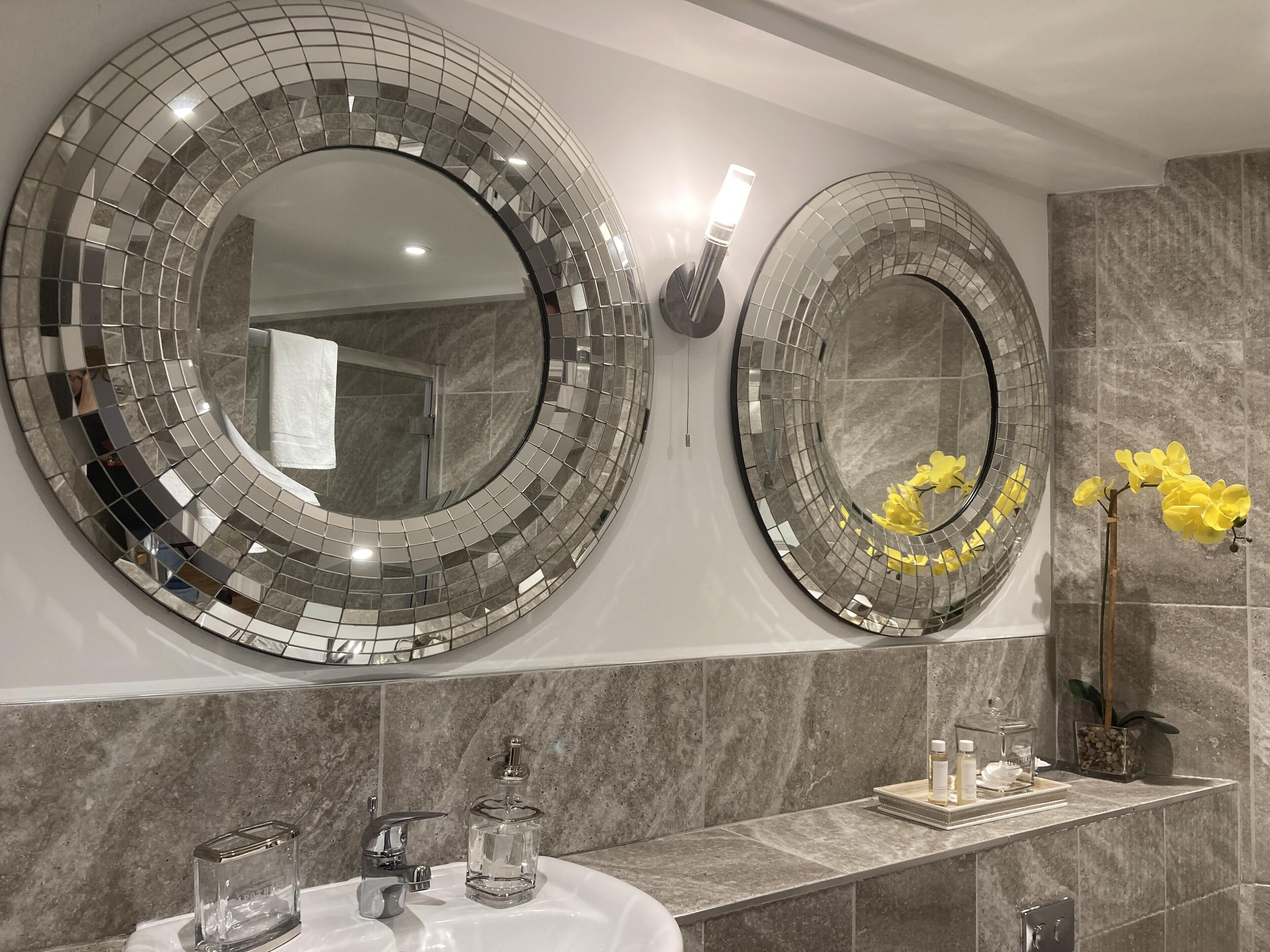 A bathroom with two large, circular mirrors with mosaic patterns, a sink with a marble countertop, and a vase with yellow flowers.