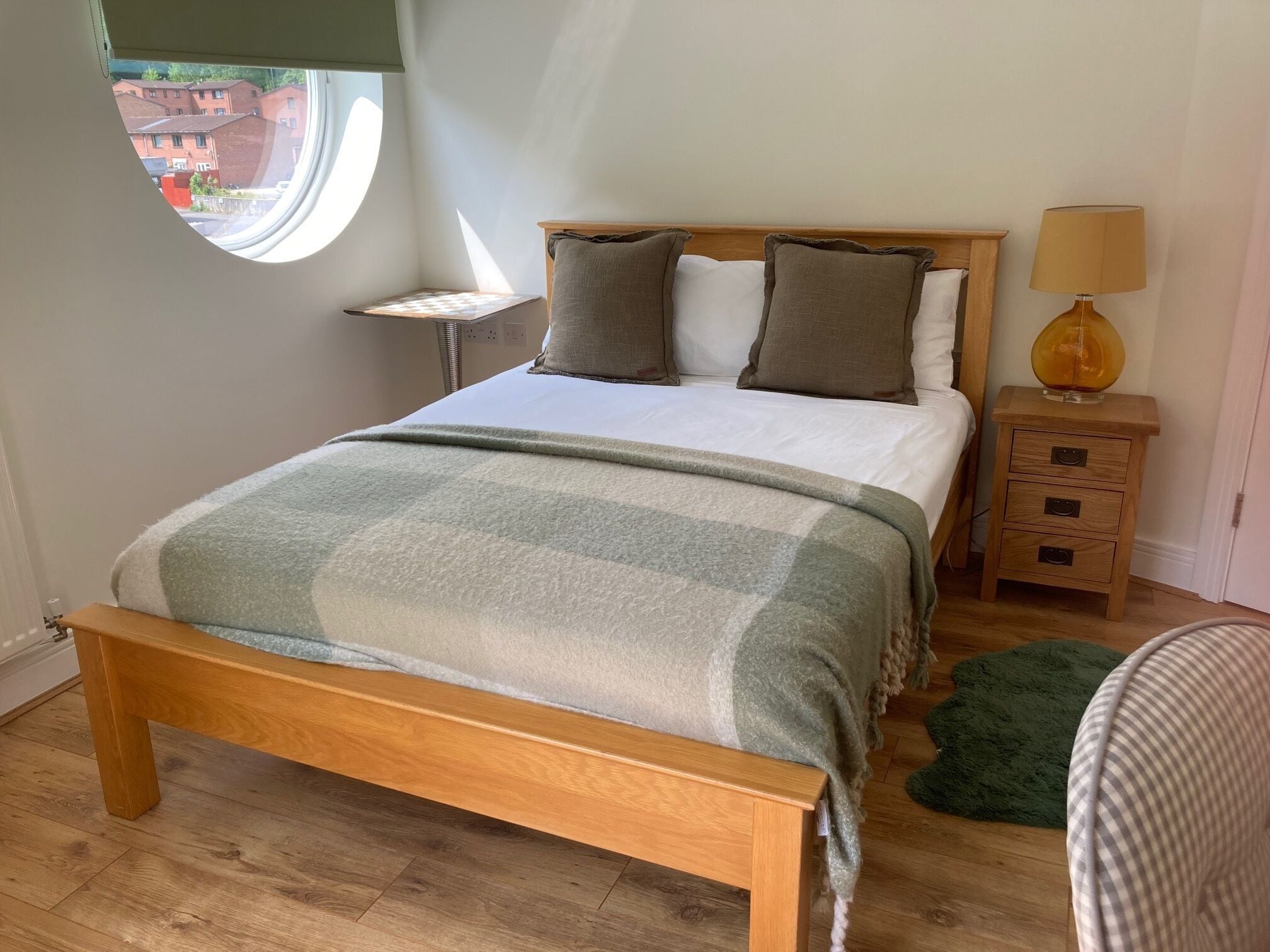A bedroom with a wooden bed, a bedside table with a lamp, a small round window, and a view of buildings outside.