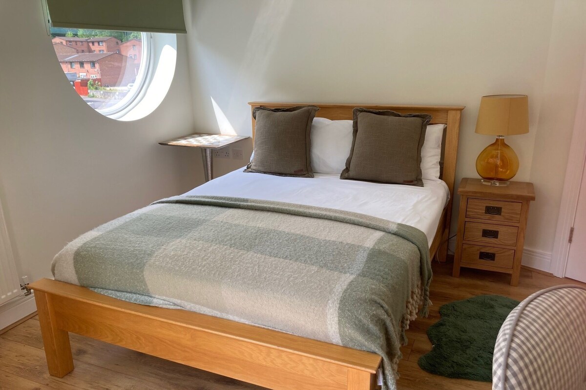 Un dormitorio con una cama de madera, una mesita de noche con lámpara, un ventanal redondo pequeño y vistas de edificios al exterior.