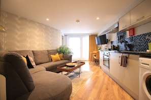 A modern living room with a sofa, coffee table, and a kitchen area with a washing machine. - Streamside Apartments (Yeovil)