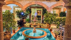 An indoor pool with a central fountain, surrounded by potted plants and stone arches.
