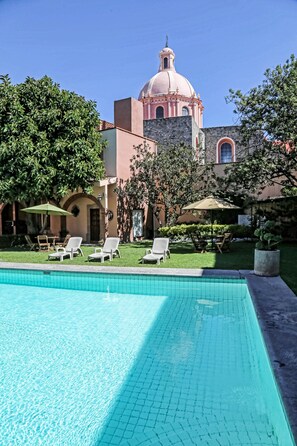 A swimming pool with lounge chairs and umbrellas, surrounded by a well-maintained lawn and a building with a pink dome and arched windows. - Hotel La Plaza de Tequisquiapan (Tequisquiapan)