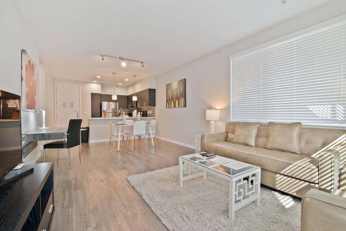 A modern living room with a beige sofa, a glass coffee table, and a dining area with a white table and chairs.