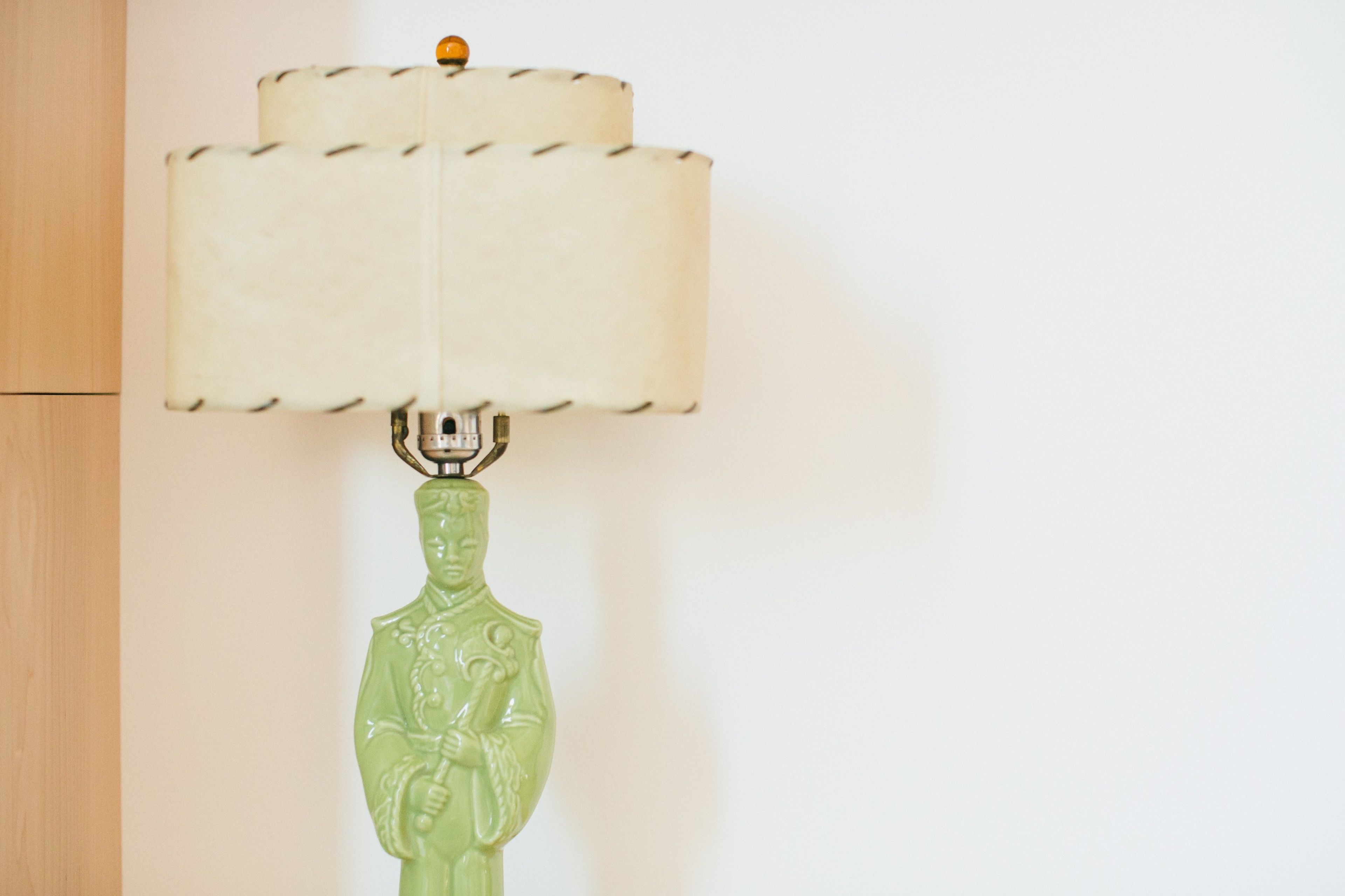 Une lampe en céramique verte, dotée d’un pied en forme de figurine et d’un abat-jour blanc à bords festonnés.