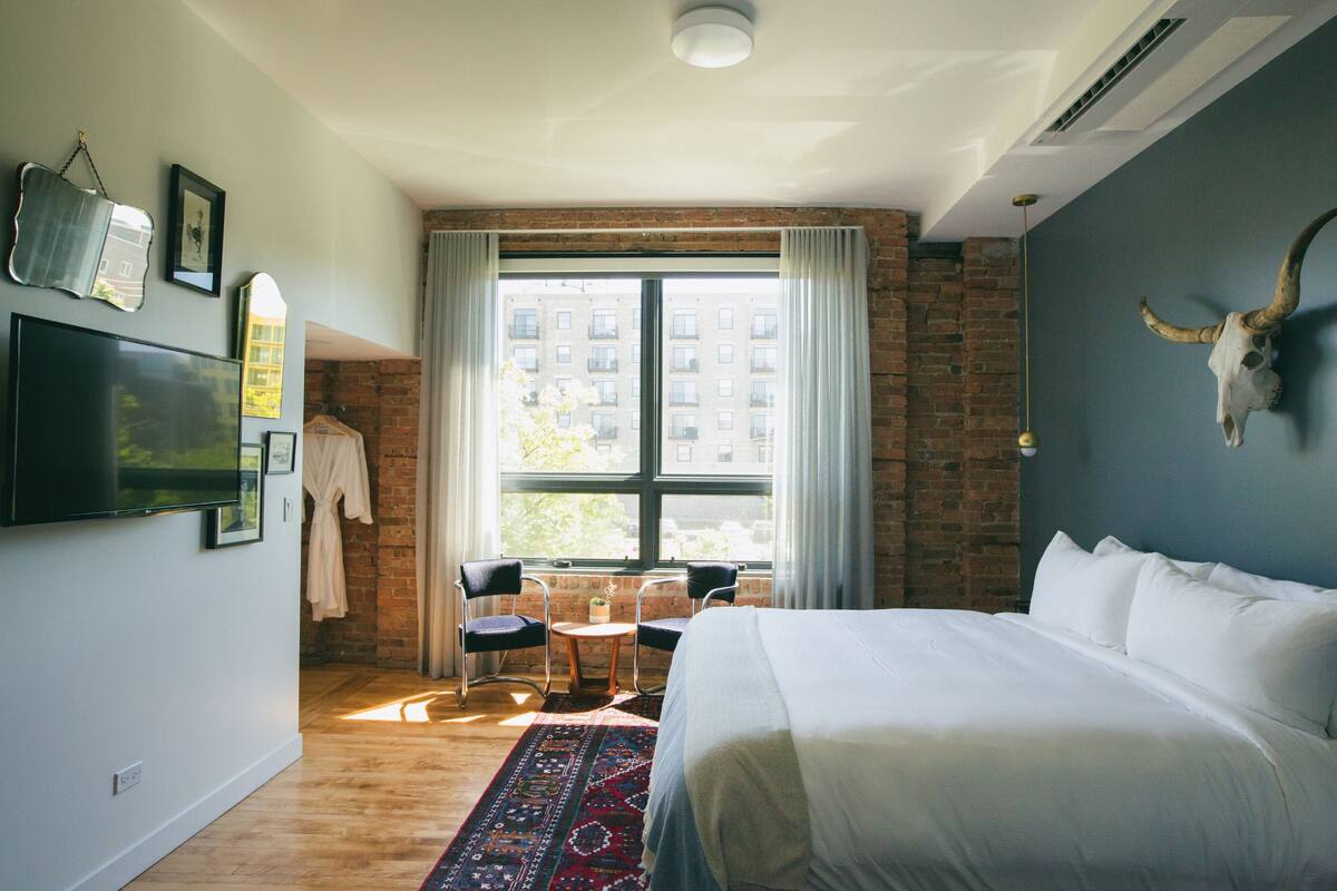 A modern hotel room with a large bed, a brick wall, a large window with curtains, a TV mounted on the wall, and a decorative bull skull on the wall.