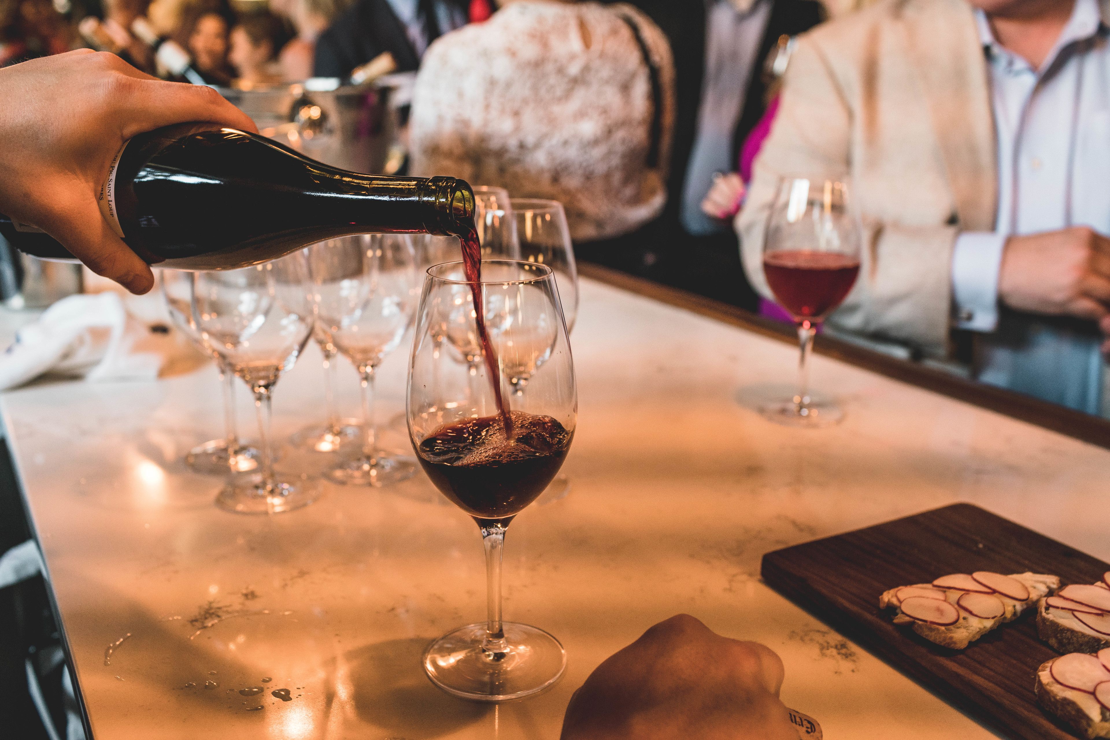 Une main versant du vin dans un verre, avec d’autres verres et une planche en bois contenant des aliments coupés en tranches en arrière-plan.