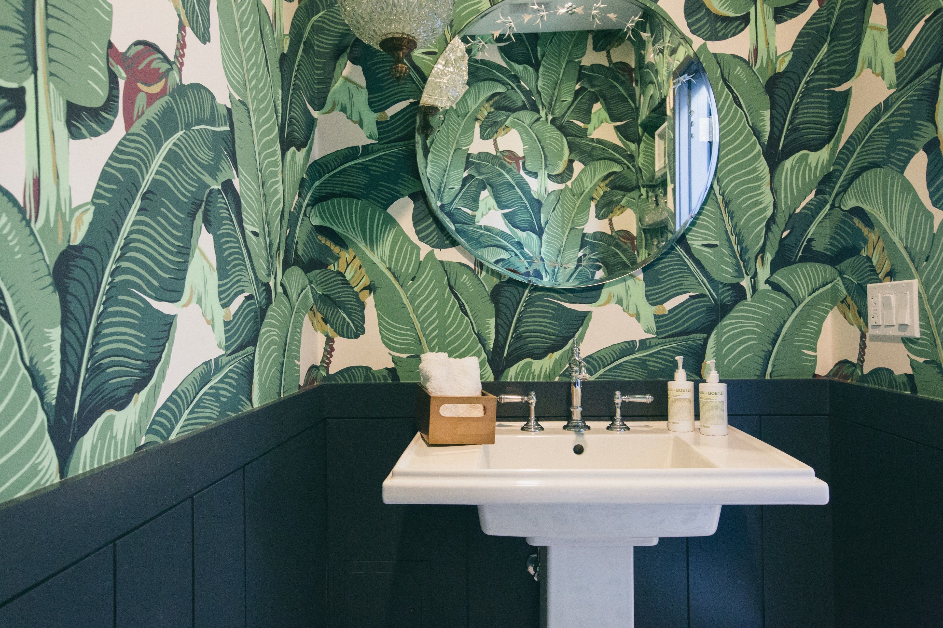 Un baño con un lavamanos blanco, un espejo y una pared con motivos de hojas tropicales.