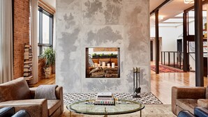 A spacious living area with a high ceiling, exposed brick walls, and a large window. A glass coffee table with books is in the center, and a fireplace with a framed picture above it is visible. - The Publishing House B&B (Chicago)