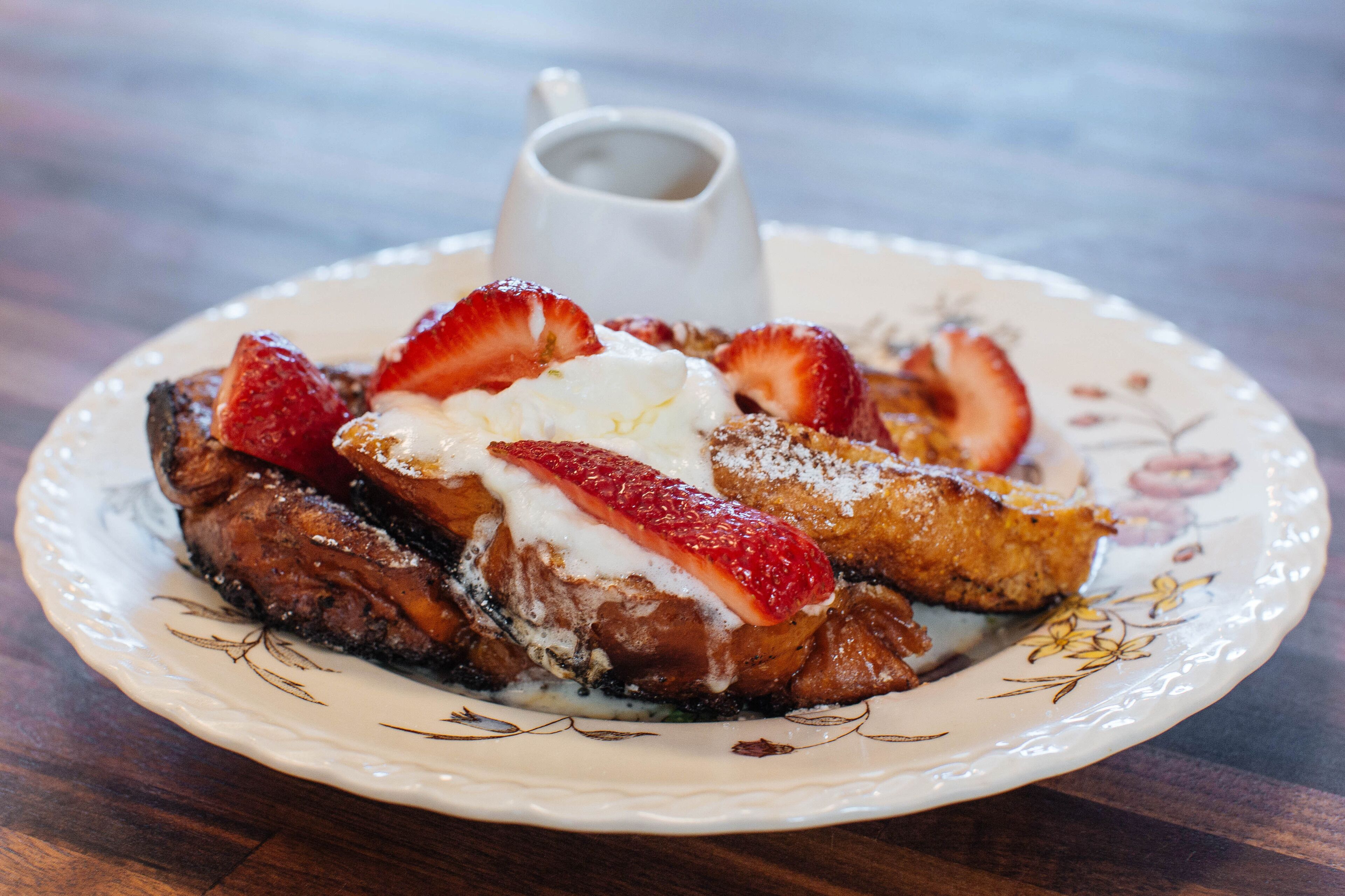 Una porción de tostadas francesas con fresas y crema batida, acompañada de una taza pequeña de almíbar.