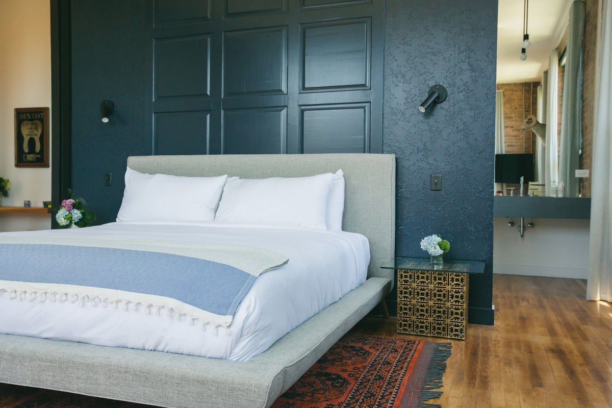 A bedroom with a large bed, dark wall paneling, a small bedside table with a vase, and a mirror.