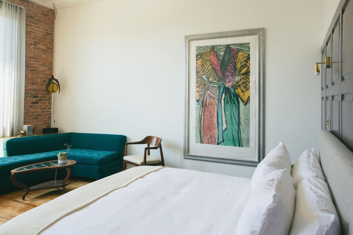 A hotel room with a bed, a teal sofa, a wooden chair, a small table, and a large abstract painting on the wall.