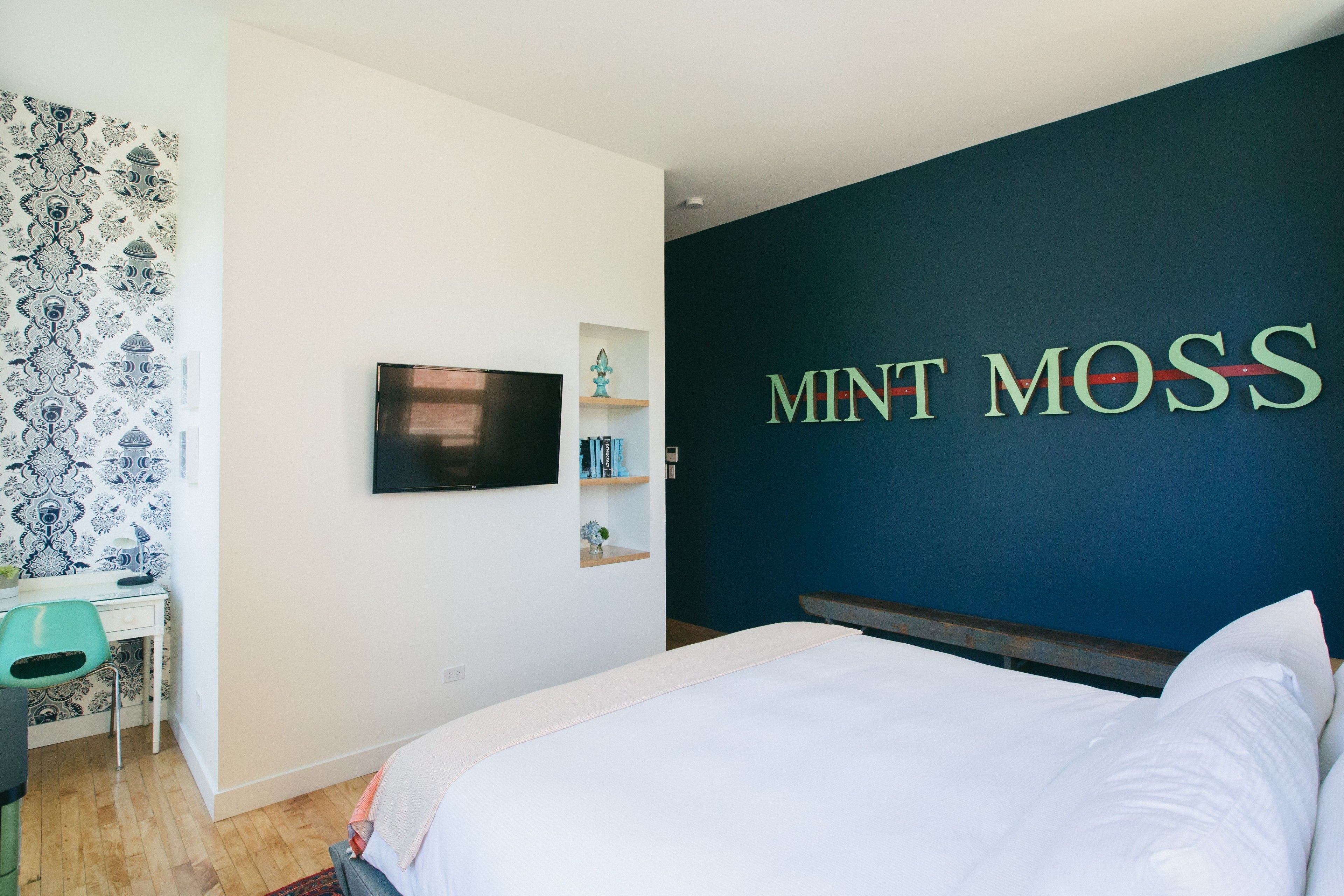 A bedroom with a bed, a desk, a chair, a TV, and a wall with the text "MINT MOSS".