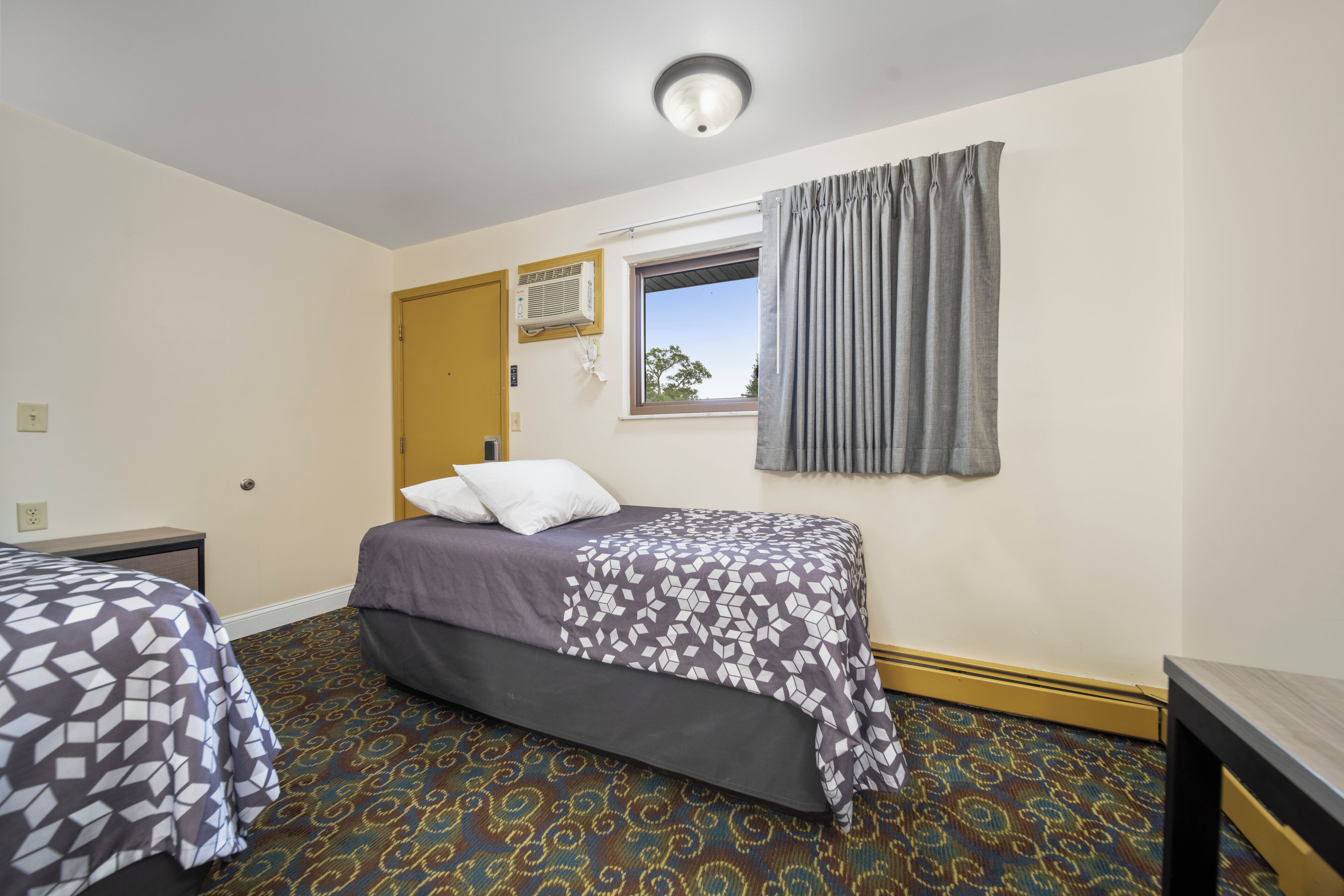 A hotel room with two beds, a window with curtains, a wall-mounted air conditioner, and a patterned carpet.