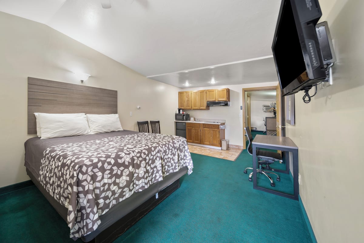 A bedroom with a bed, a desk with a chair, a kitchenette, and a TV mounted on the wall.