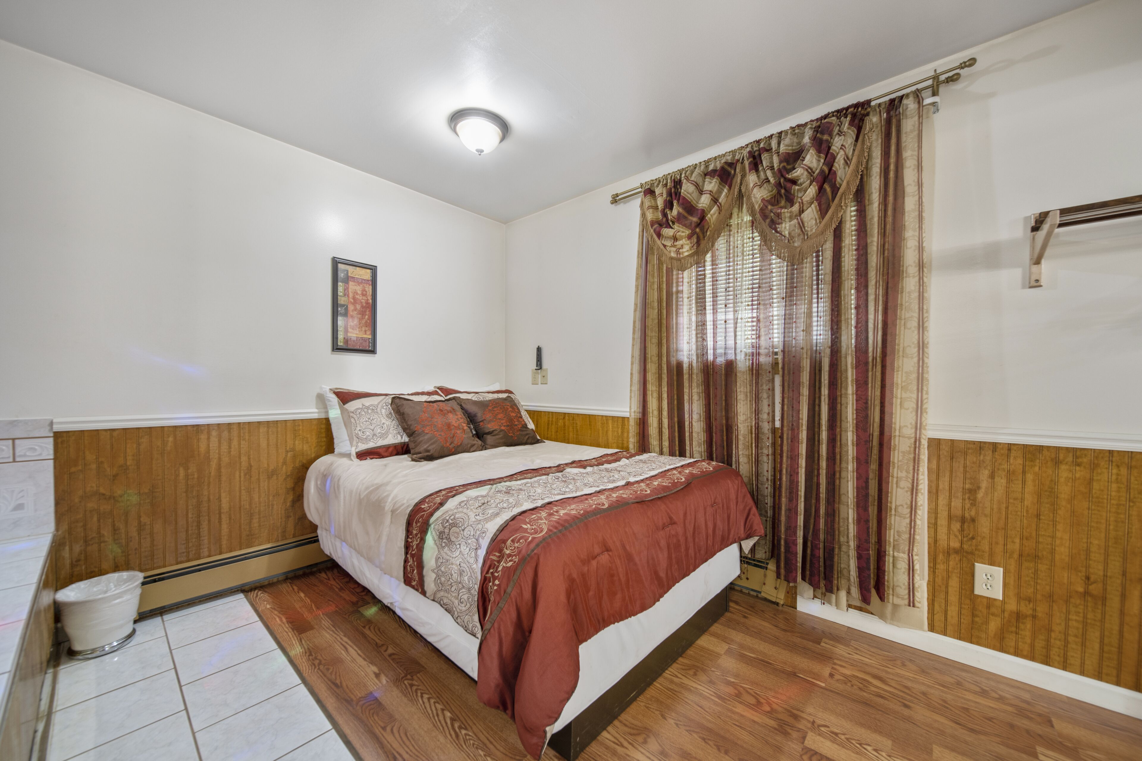 A bedroom with a wooden headboard, a bed with a patterned bedspread, a window with curtains, and a light fixture on the ceiling.