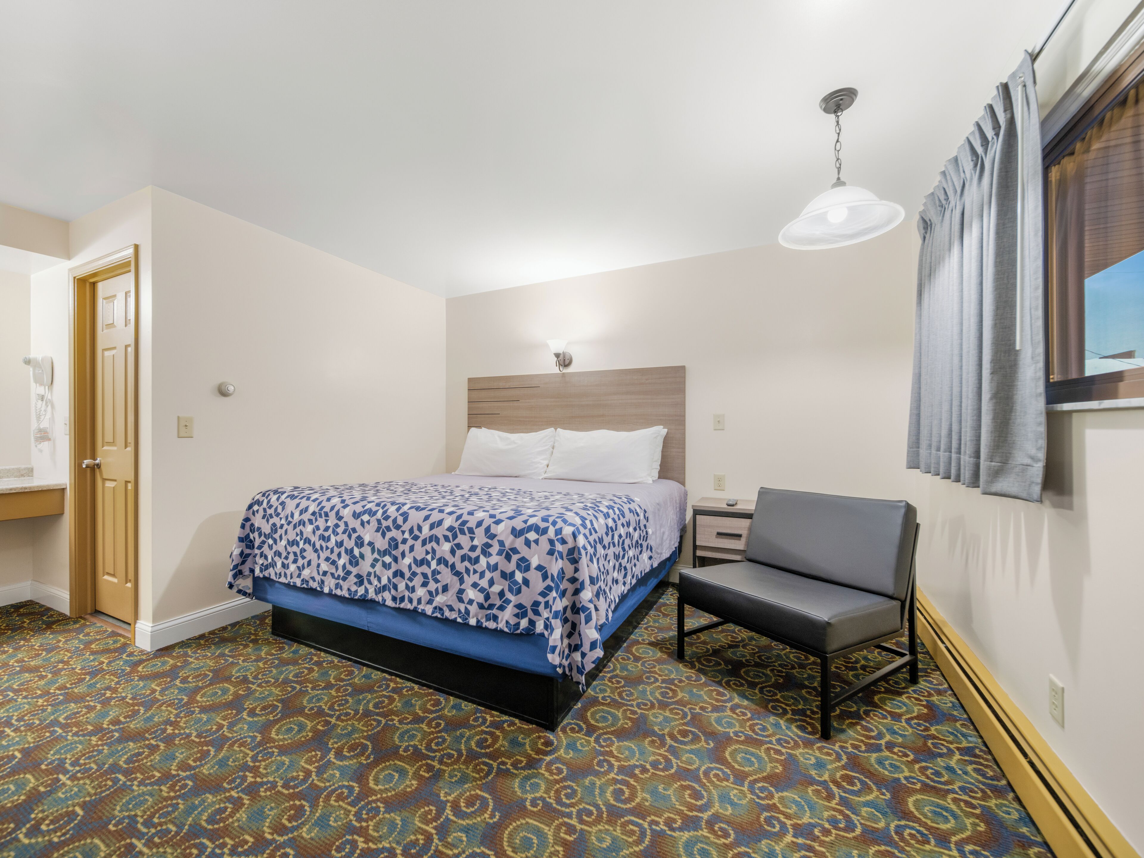 Habitación de hotel con cama, escritorio, silla, alfombra estampada, ventana con cortinas y una lámpara en el techo.