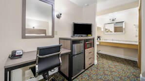 Ett hotellrum med ett skrivbord, en stol, en tv, ett kylskåp, en mikrovågsugn och ett badrum med en spegel och ett handfat.