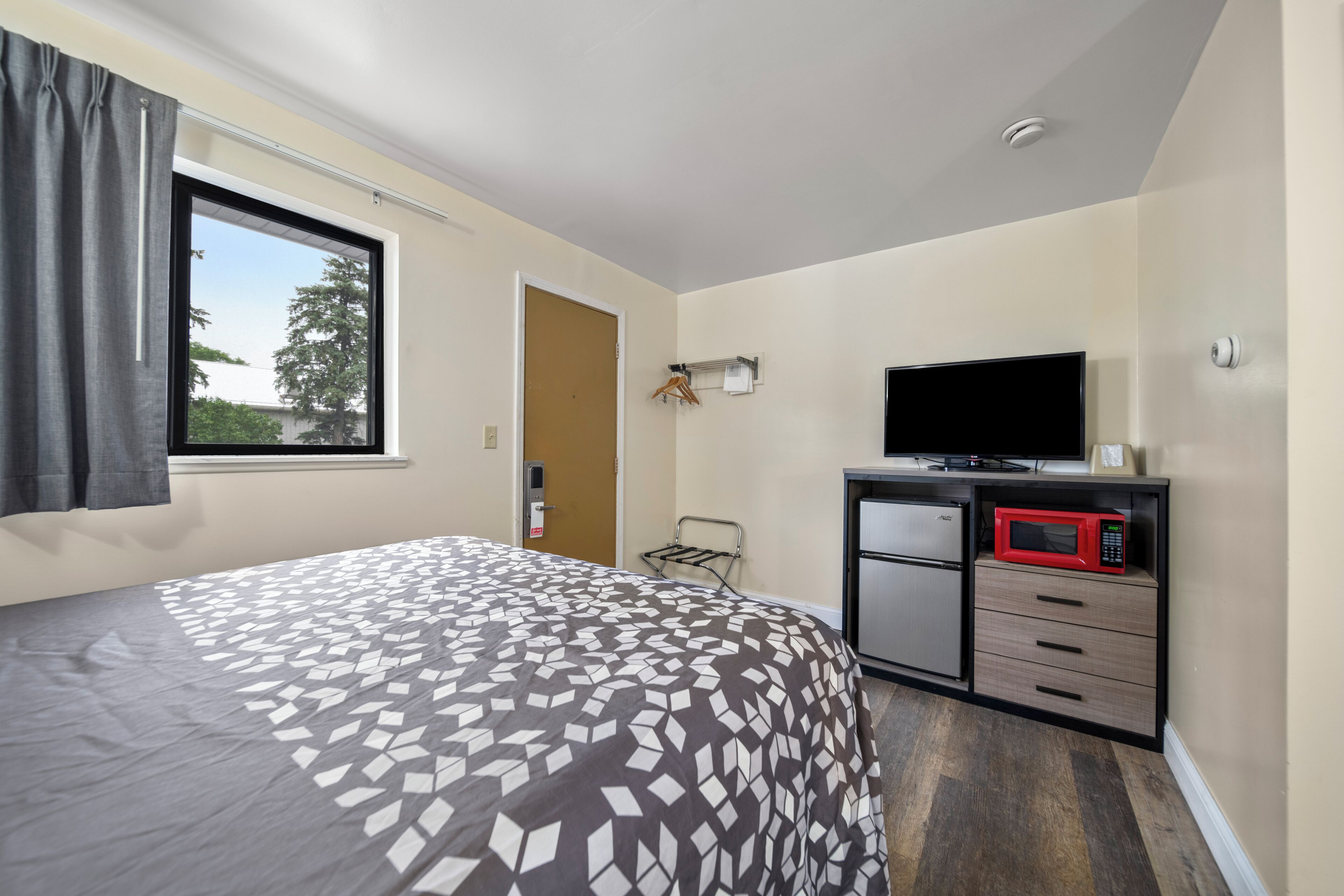 A hotel room with a bed, a desk with a microwave, a TV, and a window with curtains.