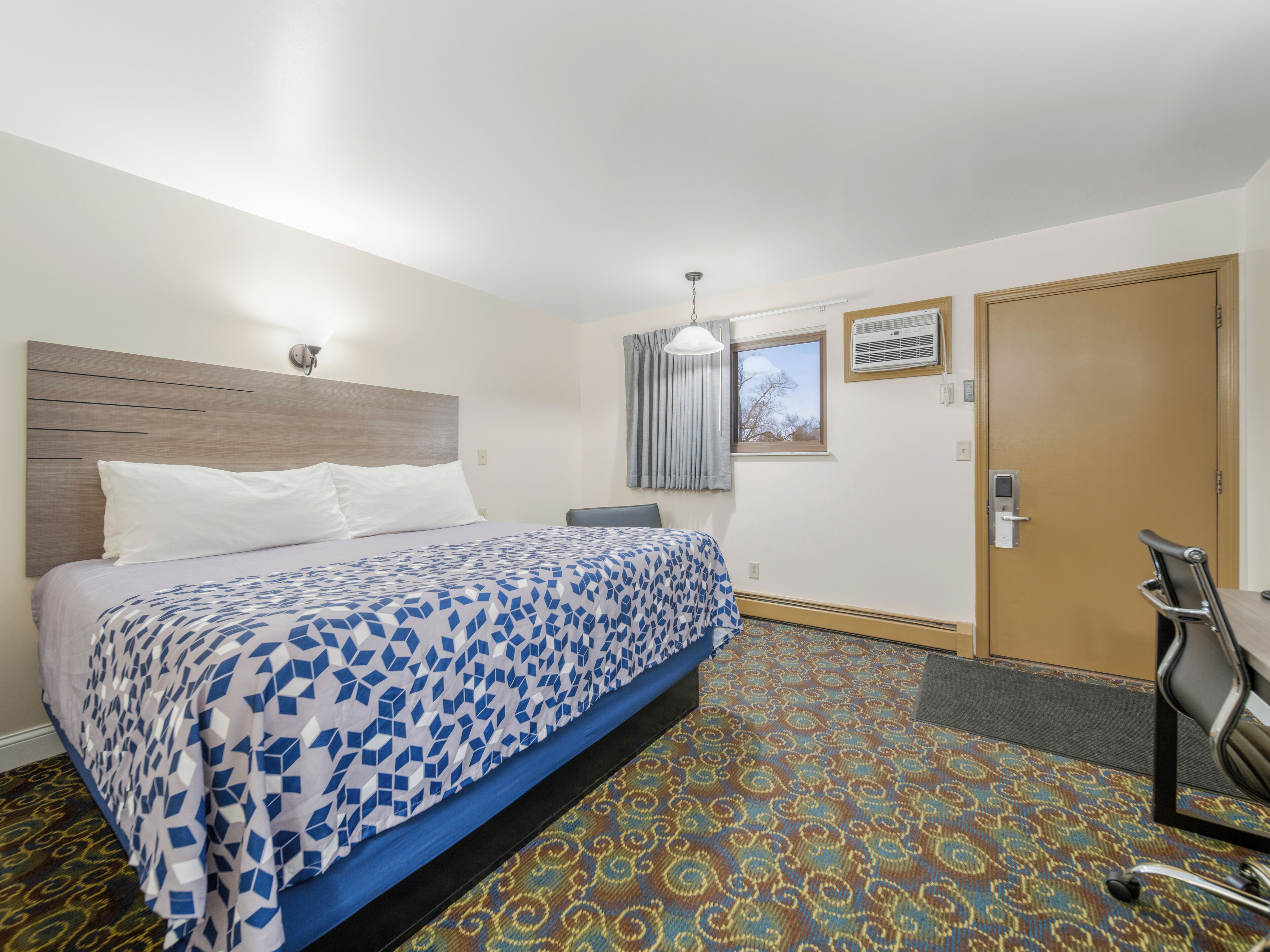 A hotel room with a bed, a desk, a chair, a window with curtains, an air conditioning unit, and a door.
