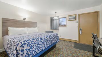 A hotel room with a bed, a desk, a chair, a window with curtains, an air conditioning unit, and a door.