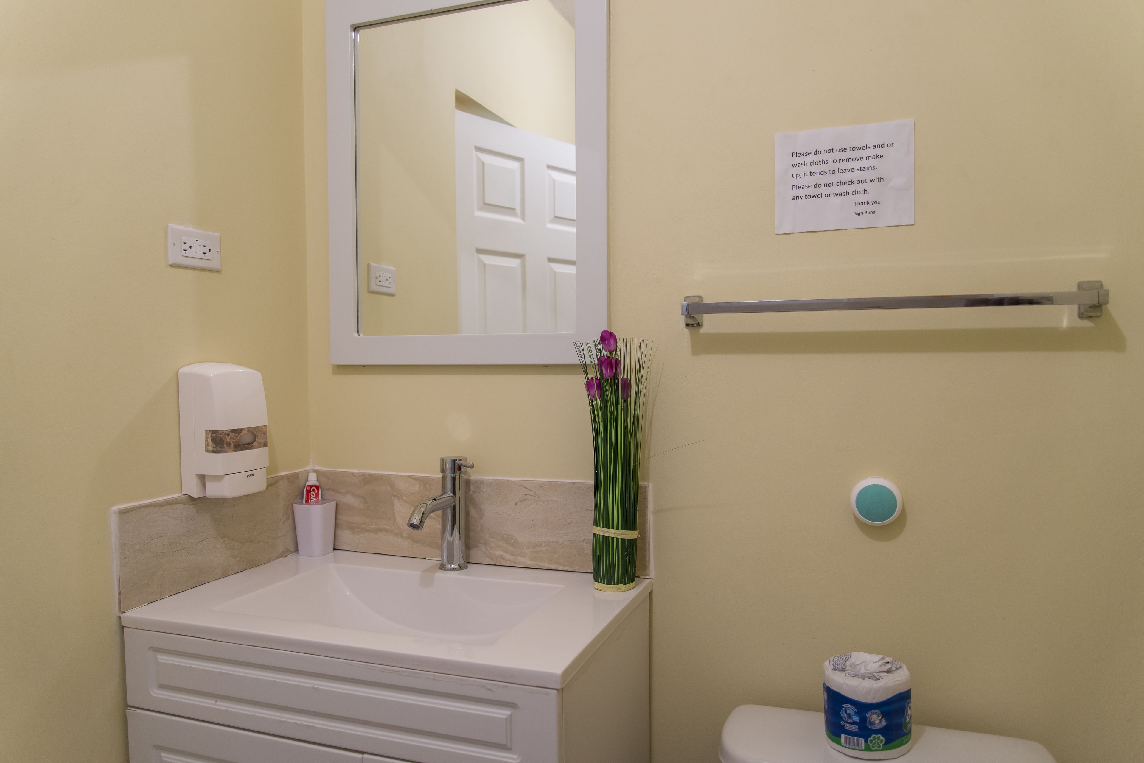 A bathroom with a sink, mirror, toilet, and a sign advising against using towels for cleaning.
