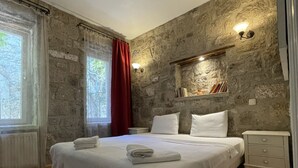 A bedroom with a large bed, stone walls, a nightstand, and a shelf with books. - Kallista Alaçatı (Cesme)