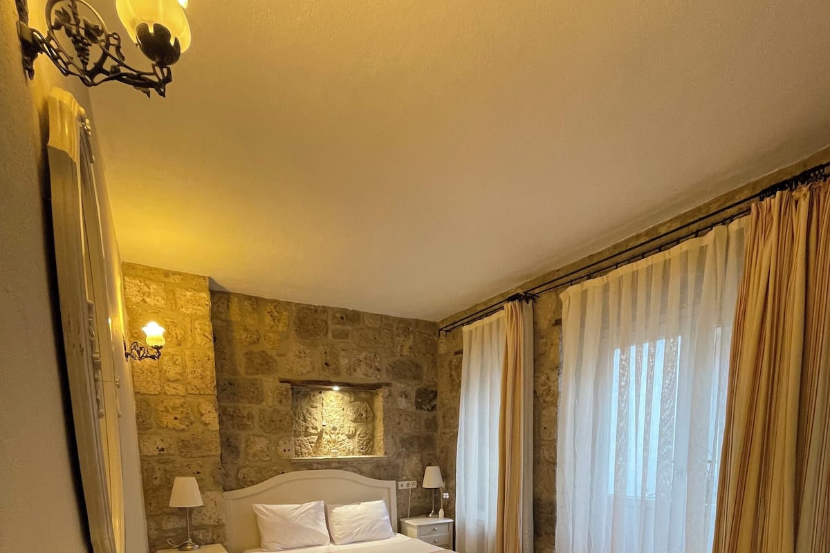 A bedroom with a bed, a desk, and a chair. There is a stone wall and a window with curtains.