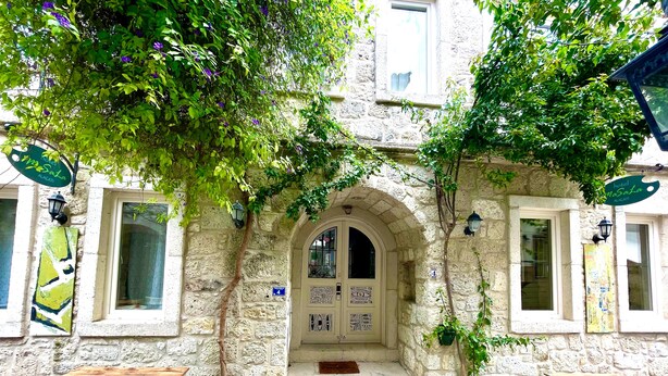 A stone building with a central arched entrance, two windows on either side, and a door with a sign. There are two tables and a bench outside. - Kallista Alaçatı (Cesme)