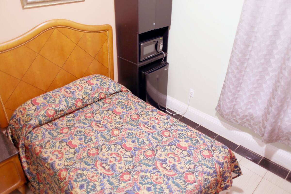 A bed with a patterned bedspread, a wooden headboard, a black microwave, and a white power outlet on the wall.