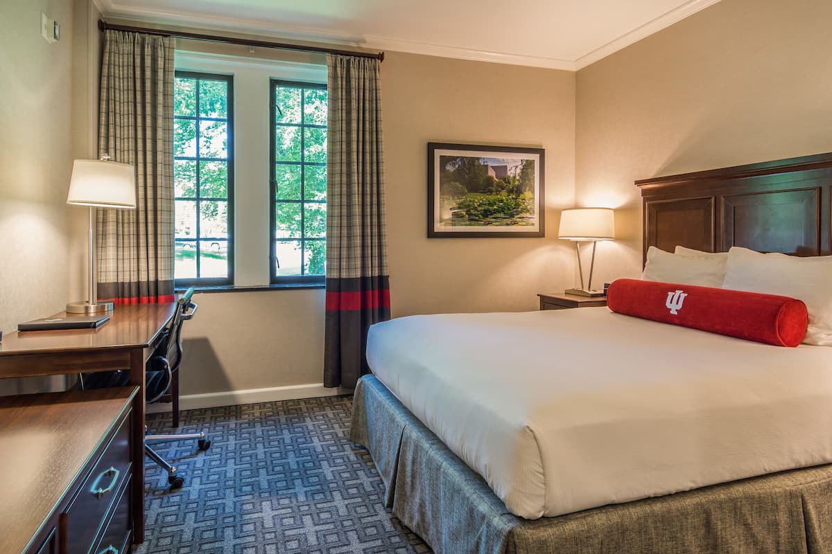 A hotel room with a bed, desk, and chair, a window with curtains, a framed picture on the wall, and a red pillow on the bed.