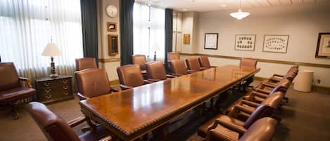 Un salón de conferencias con una gran mesa de madera rodeada de sillones de cuero, un reloj en la pared y una ventana con cortinas.
