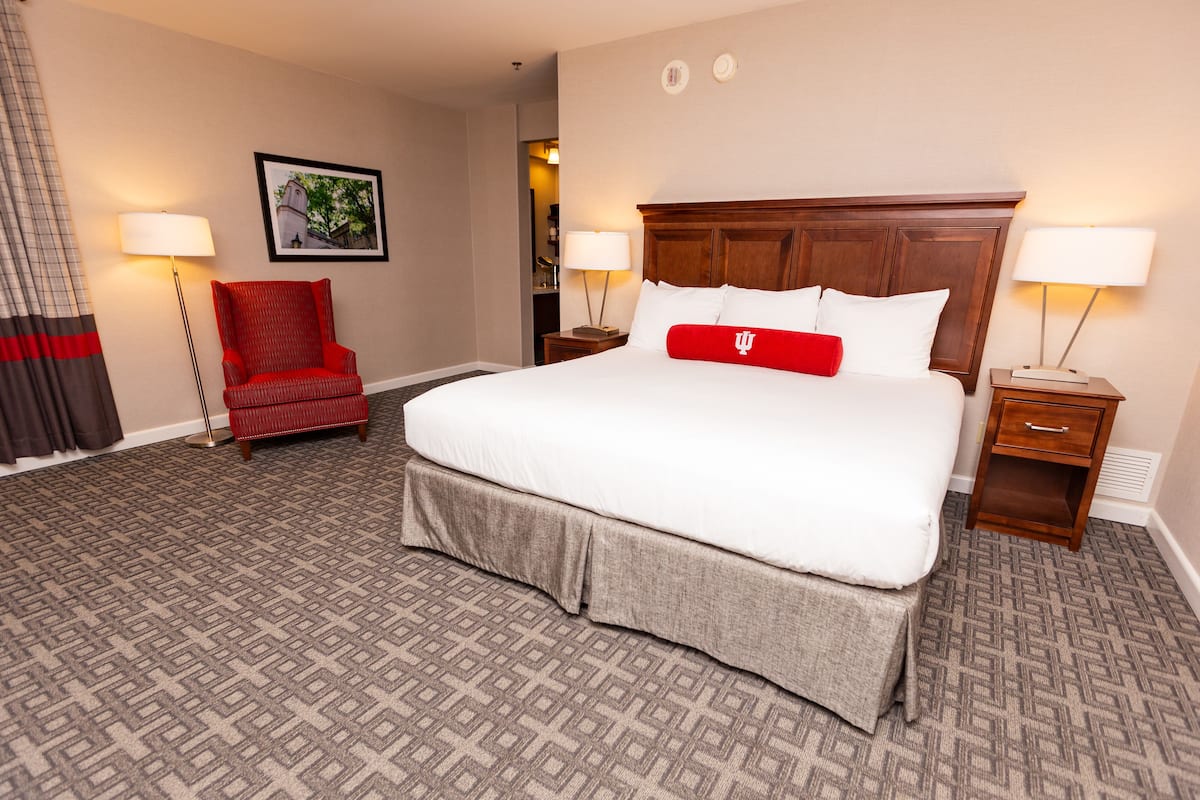 A hotel room with a large bed, a red chair, two bedside tables with lamps, a framed picture on the wall, and a patterned carpet.