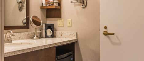 A bathroom with a sink, mirror, and a coffee maker. There is a shelf with toiletries and a towel rack.