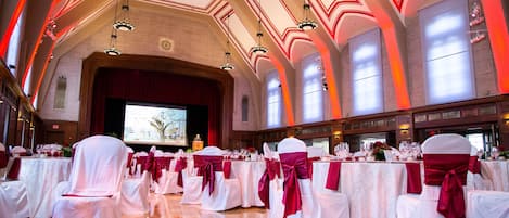 Un salón de banquetes con mesas y sillas blancas, cintas rojas y un escenario con una pantalla grande.