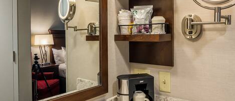 A bathroom with a large mirror, a sink, a coffee maker, and a shelf with toiletries.
