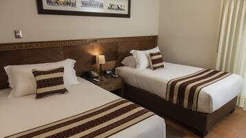 A hotel room with two beds, each with patterned bedding and pillows, a nightstand with a lamp, and a framed picture above the beds.