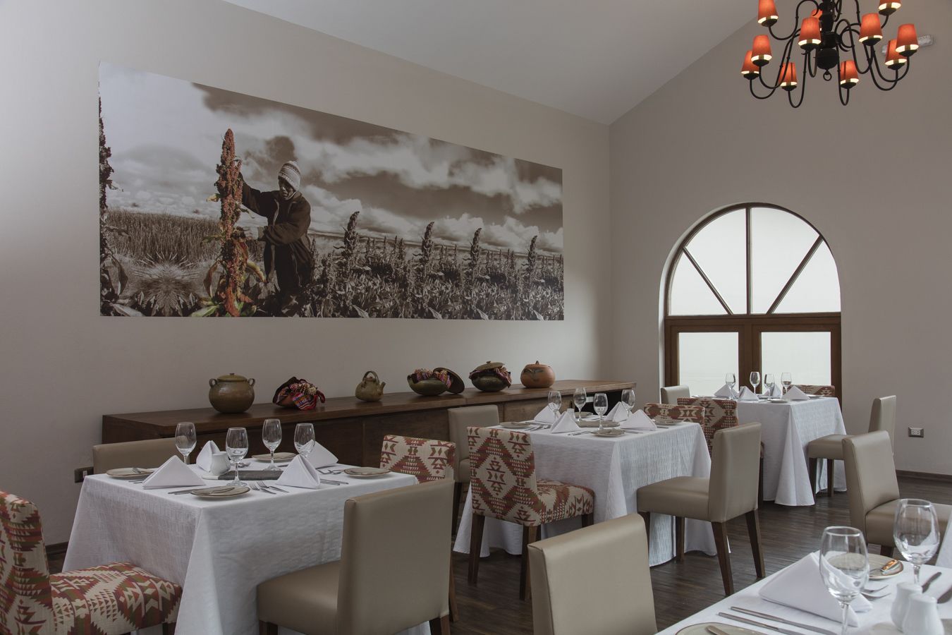 Uma sala de jantar com mesas preparadas para quatro pessoas, um quadro grande na parede e um candelabro.