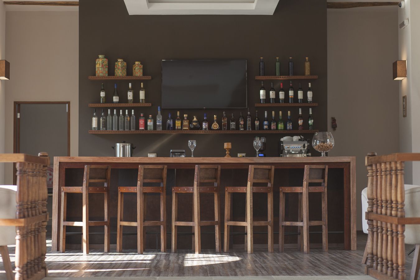 Zona de bar con taburetes de madera, un televisor sobre la barra y estantes con botellas.