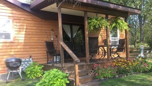 Una casa de madera con porche cubierto, mobiliario exterior, una parrilla y un jardín con flores.