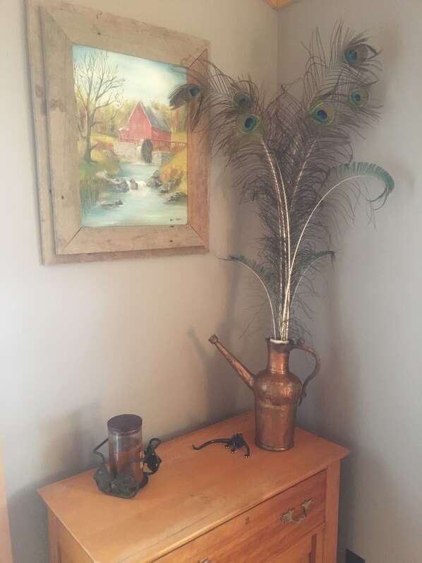 A wooden dresser with a copper vase holding peacock feathers, a painting of a rural scene, and a small glass container on the dresser. - Mill Pond Hideaway (Luther)