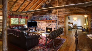 Ein Interieur einer Blockhütte mit Kamin, Ledermöbeln, einem Fernseher und einem Hirschgeweih, das an der Decke befestigt ist.