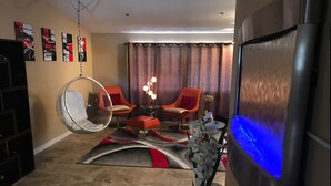 Ein modernes Wohnzimmer mit einem großen Flachbildfernseher, zwei orangefarbenen Sesseln, einem runden Spiegel und einer Zierpflanze.